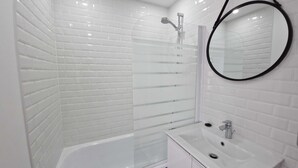 Een moderne badkamer met witte metro tegels, een douche met glazen wand, een witte badkuip en een ronde spiegel met een zwarte lijst.
