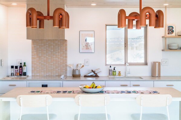 A modern kitchen with a large island, pendant lights, a window with a view, and various kitchen items. - Stylish, Secluded, Peaceful Home with Hot Tub, EV Charger, Star Gazing (Yucca Valley)