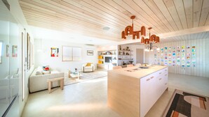 A modern living room with a central island, a sofa, a coffee table, and a dining area with a large island. There are shelves with decorative items and a wall with colorful artwork. - Stylish, Secluded, Peaceful Home with Hot Tub, EV Charger, Star Gazing (Yucca Valley)