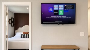 A modern hotel room with a bed, a bedside table, and a mounted TV displaying streaming apps.