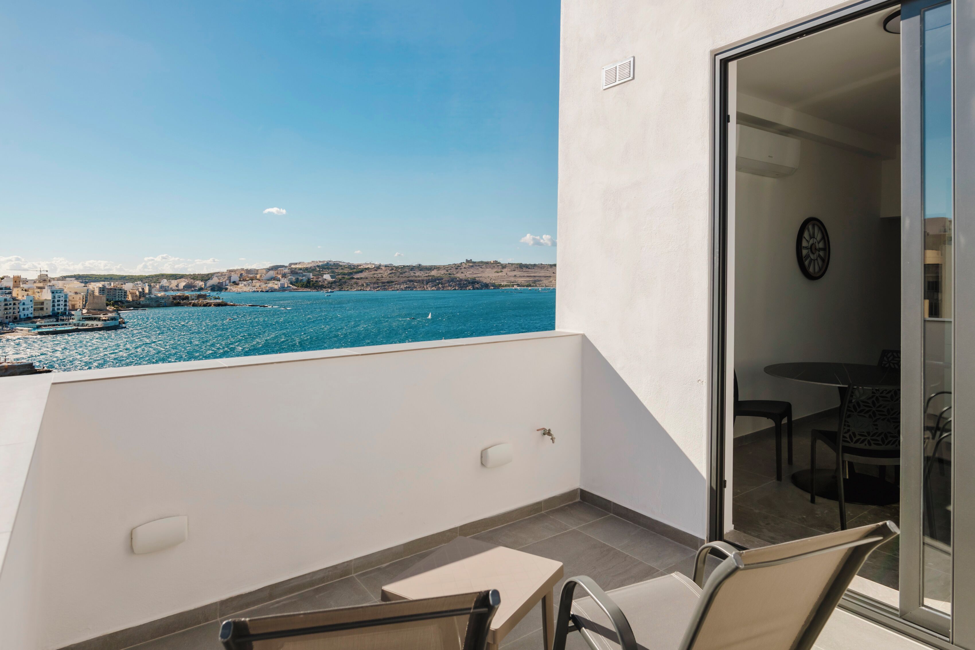 Een balkon met uitzicht op zee en gebouwen, een tafel met stoelen, en een deur die naar een kamer leidt met een klok.