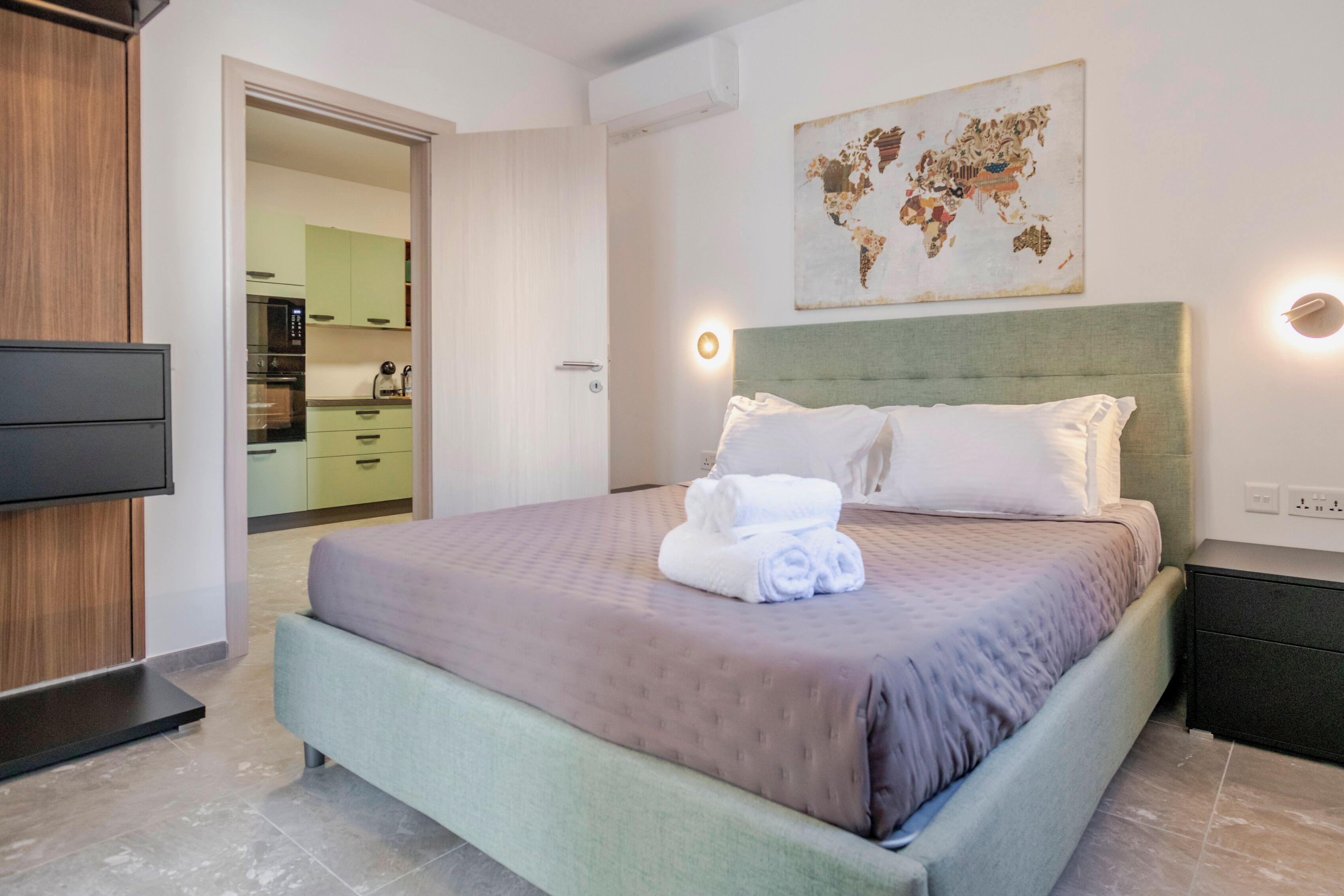 Habitación de hotel moderna con una cama grande, un televisor de pantalla plana, una obra de arte con un mapa mundial y vista al área de la cocina.
