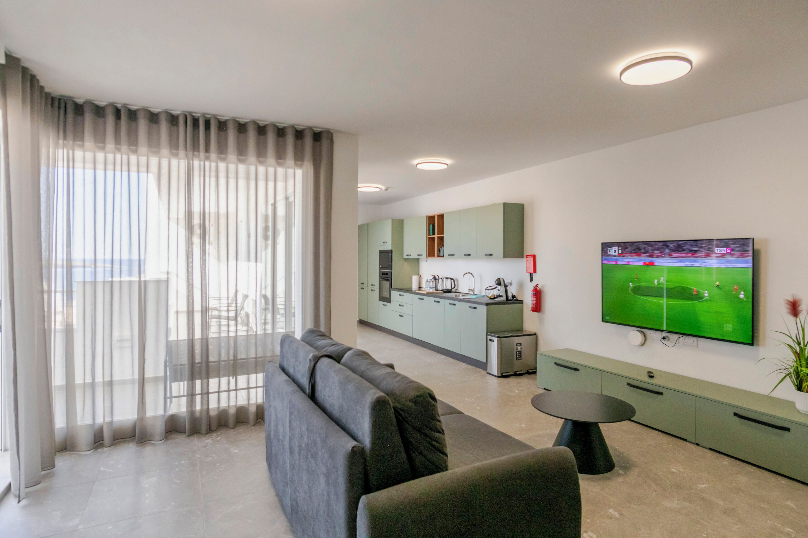 Una sala de estar moderna con un sofá gris, una mesita redonda y una cocina al fondo.