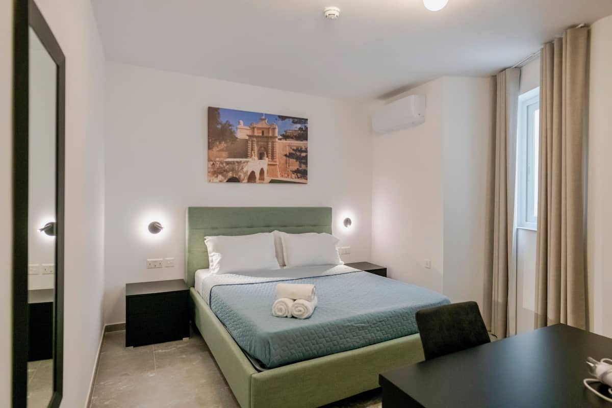 Une chambre d’hôtel comprenant un lit, des tables de chevet, un bureau, une chaise, une fenêtre avec des rideaux et un tableau accroché au mur.