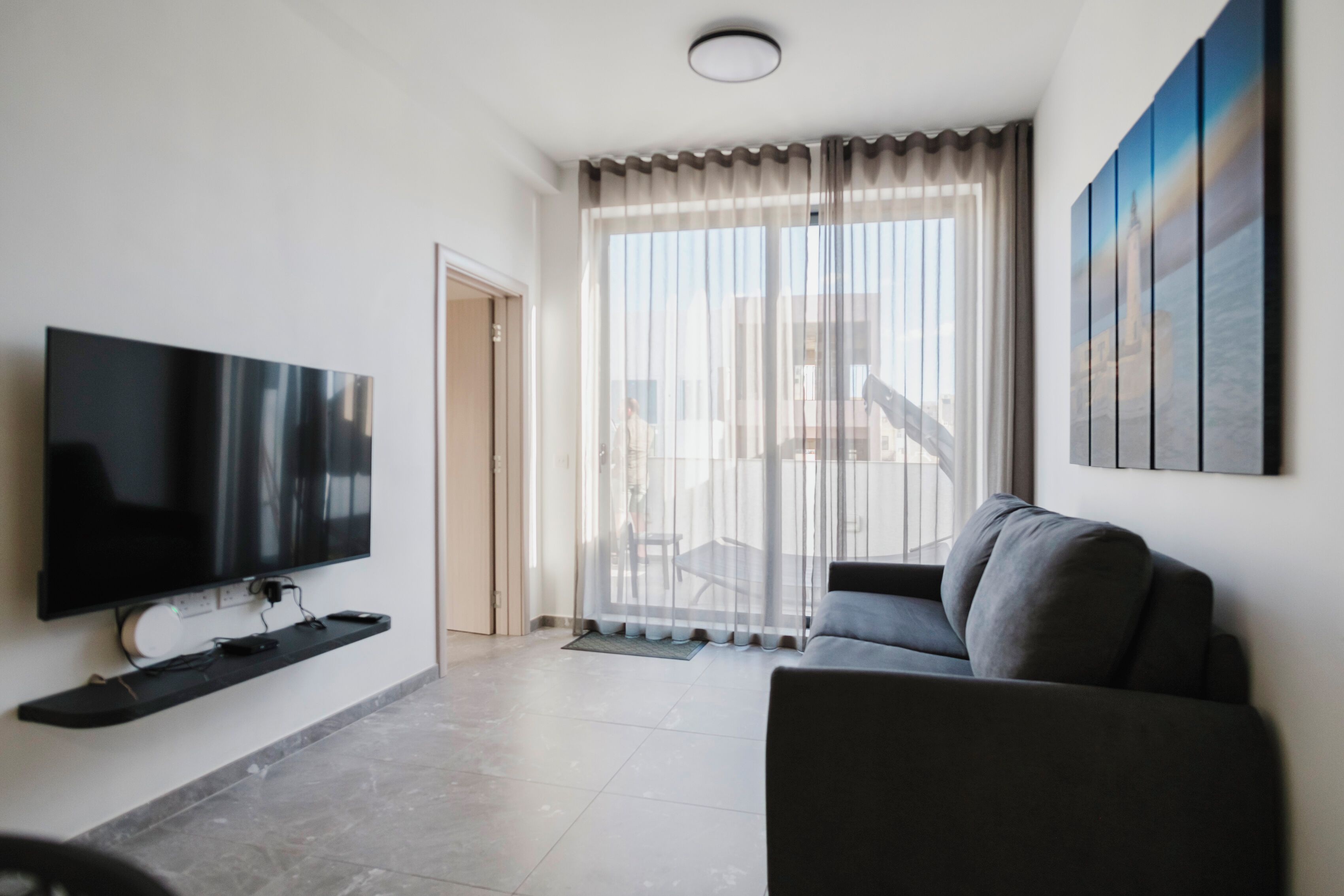 Un salon moderne avec un téléviseur à écran plat, un canapé gris et une grande fenêtre ornée de rideaux légers.