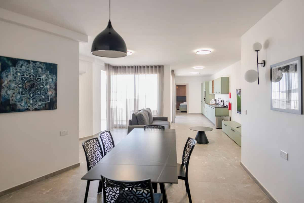 Un espace repas moderne avec une longue table et des chaises, un luminaire suspendu et une vue sur la cuisine.