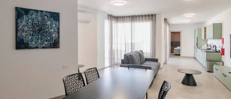 Una cocina moderna con armarios verdes, un comedor con una mesa larga y sillas, y una sala de estar con sofá y cortinas.