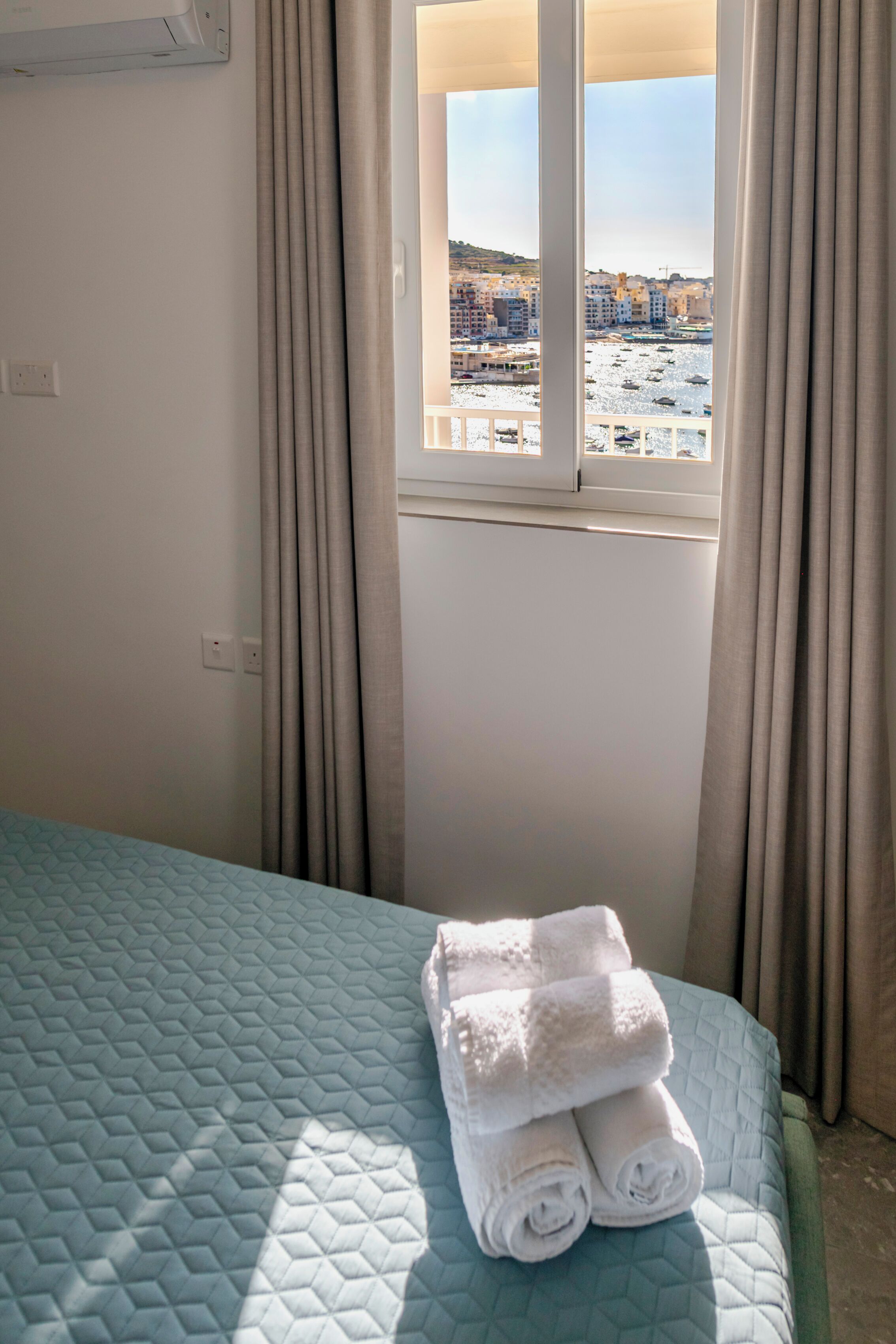 Una habitación de hotel con cama, ventana con cortinas y vista a una playa y edificios.
