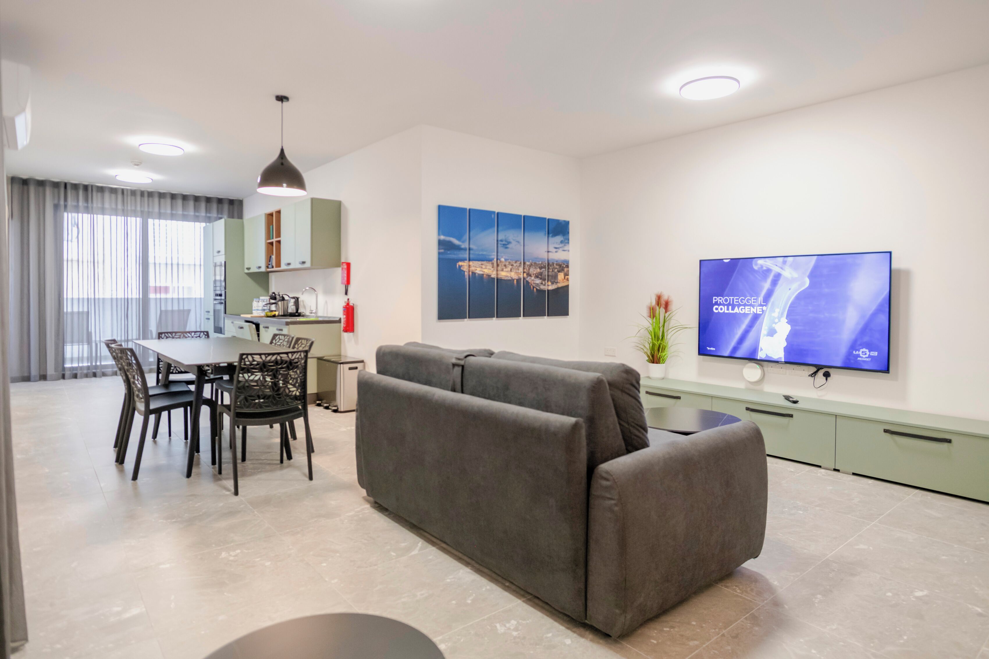 Una sala de estar moderna con un sofá gris, una mesa de comedor con sillas, un televisor de pantalla plana montado en la pared y una obra de arte con un paisaje urbano.