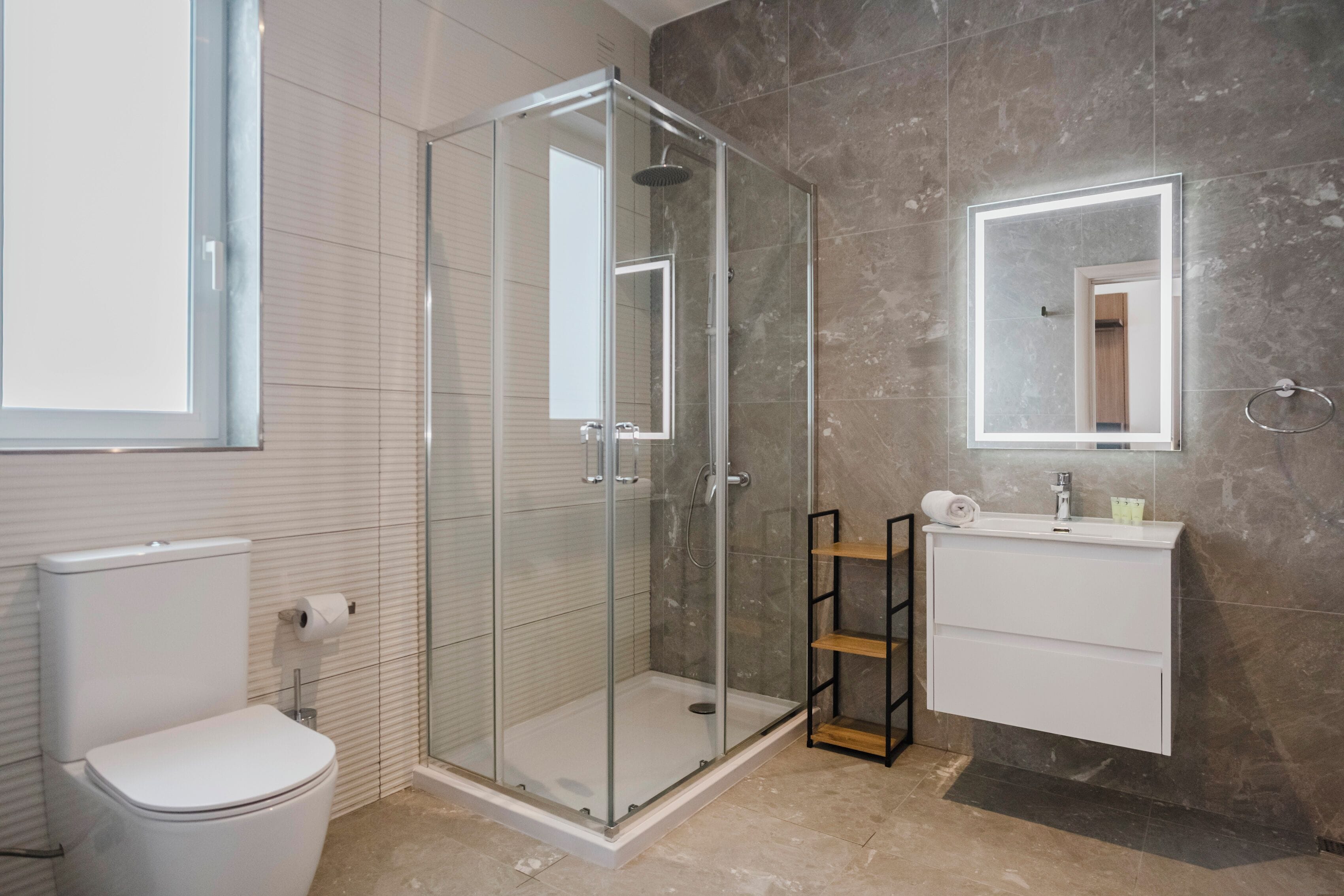 A modern bathroom with a glass-enclosed shower, a toilet, a sink with a mirror, and a shelf with toiletries.