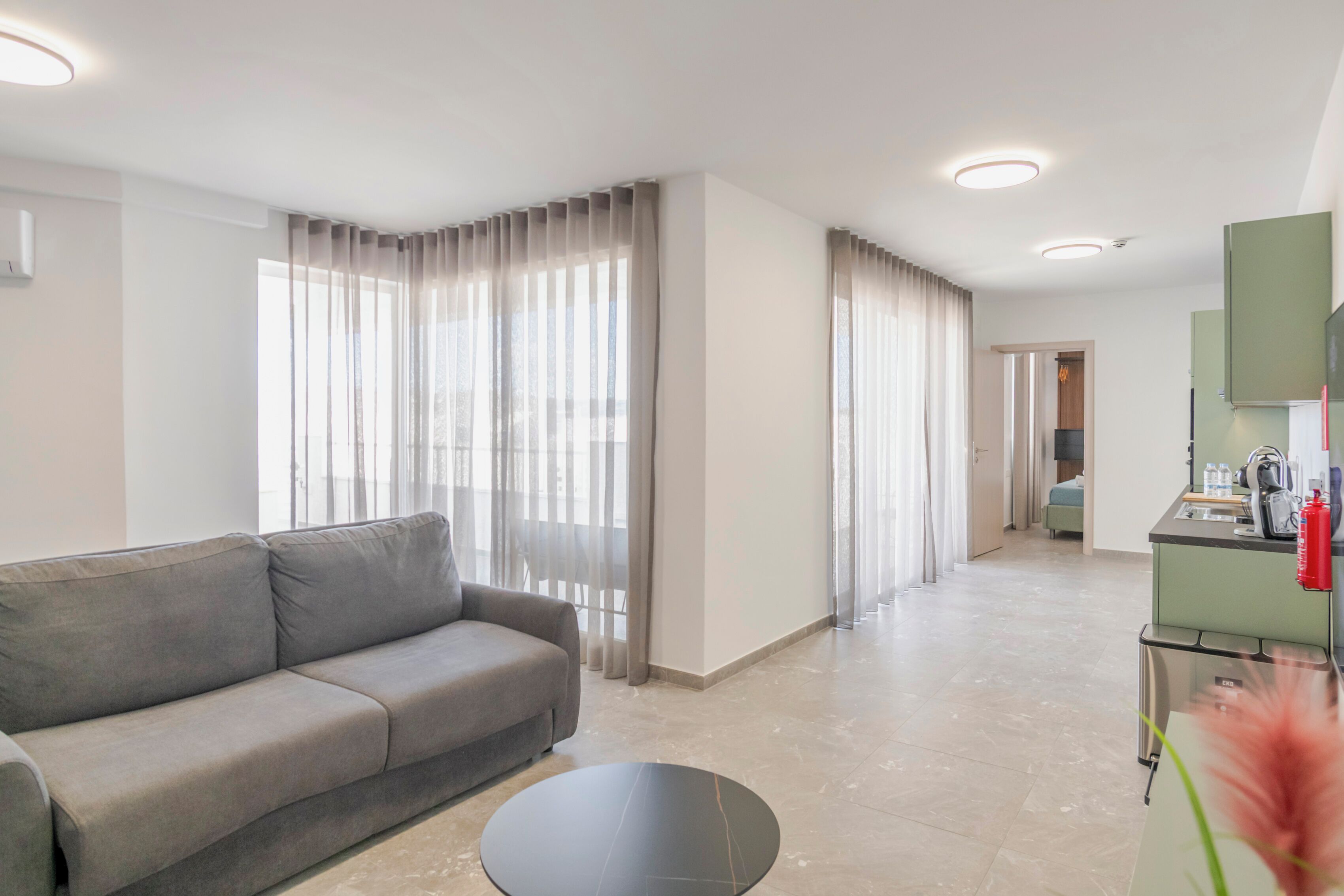 A modern living room with a grey sofa and a round coffee table, a kitchen area with a green countertop, and a view through the curtains.