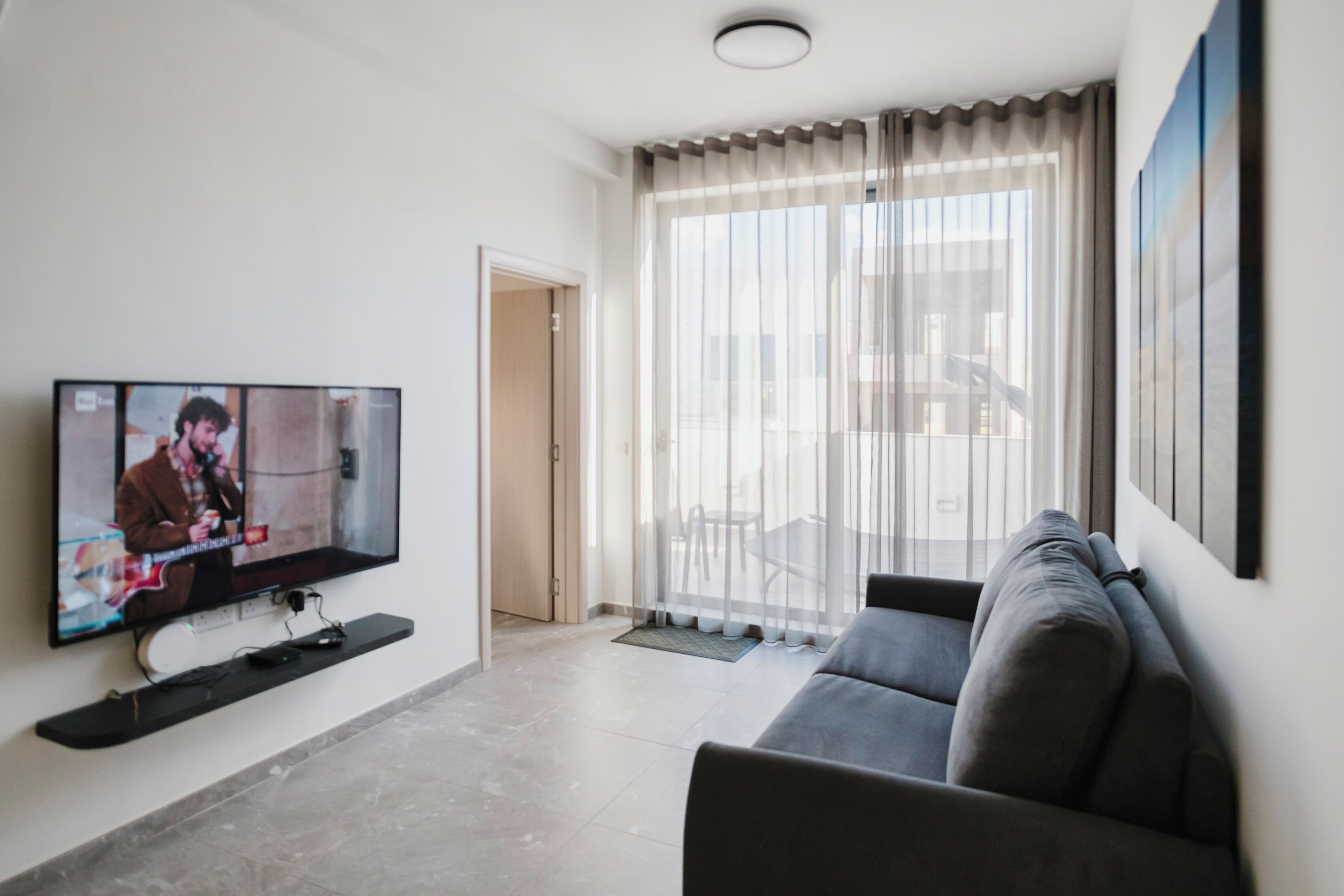A modern living room with a flat-screen TV mounted on the wall, a grey sofa, and a large window with sheer curtains.