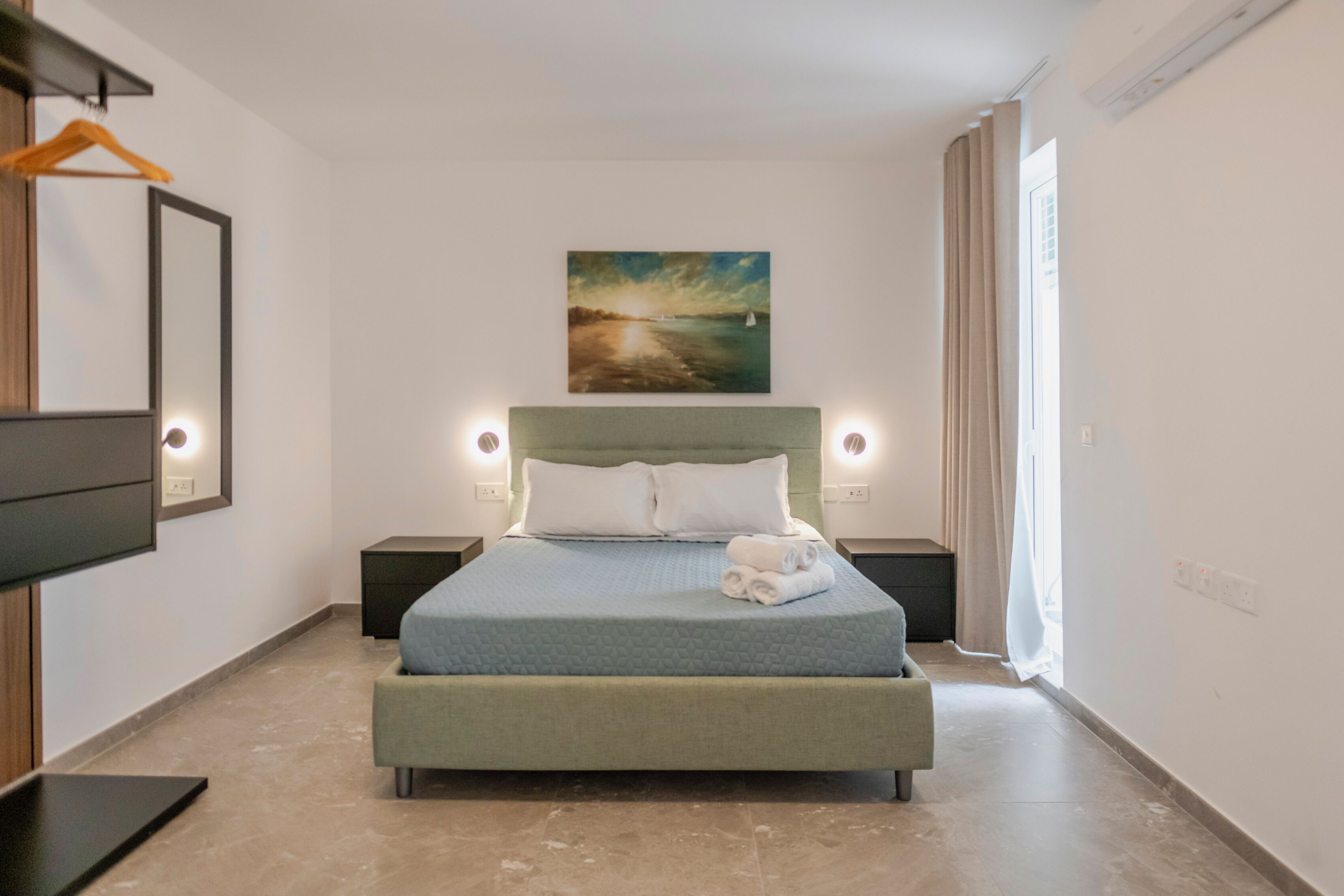 Une chambre à coucher bien rangée, avec un lit placé au centre, flanqué de tables de chevet sur lesquelles reposent des lampes, un tableau accroché au mur et une fenêtre ornée de rideaux.