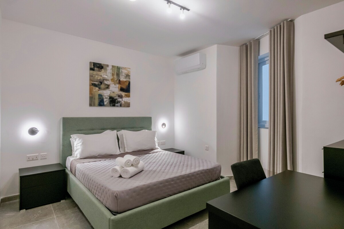 A neatly arranged bedroom with a bed, bedside tables, a desk, and a window with curtains.
