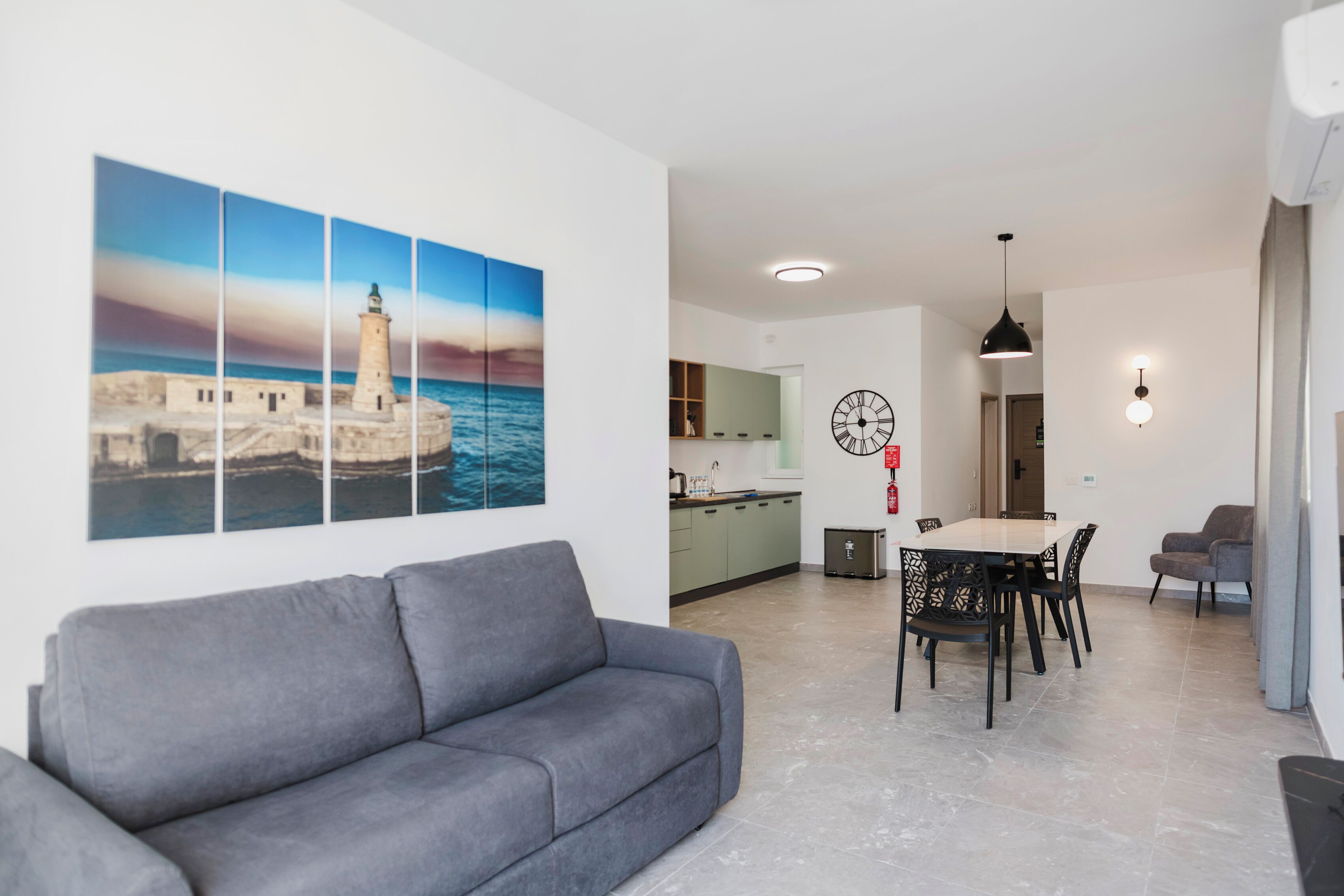 Una sala de estar moderna con un sofá gris, una zona de comedor con una mesa y sillas, una cocina al fondo y un cuadro grande con una escena costera en la pared.