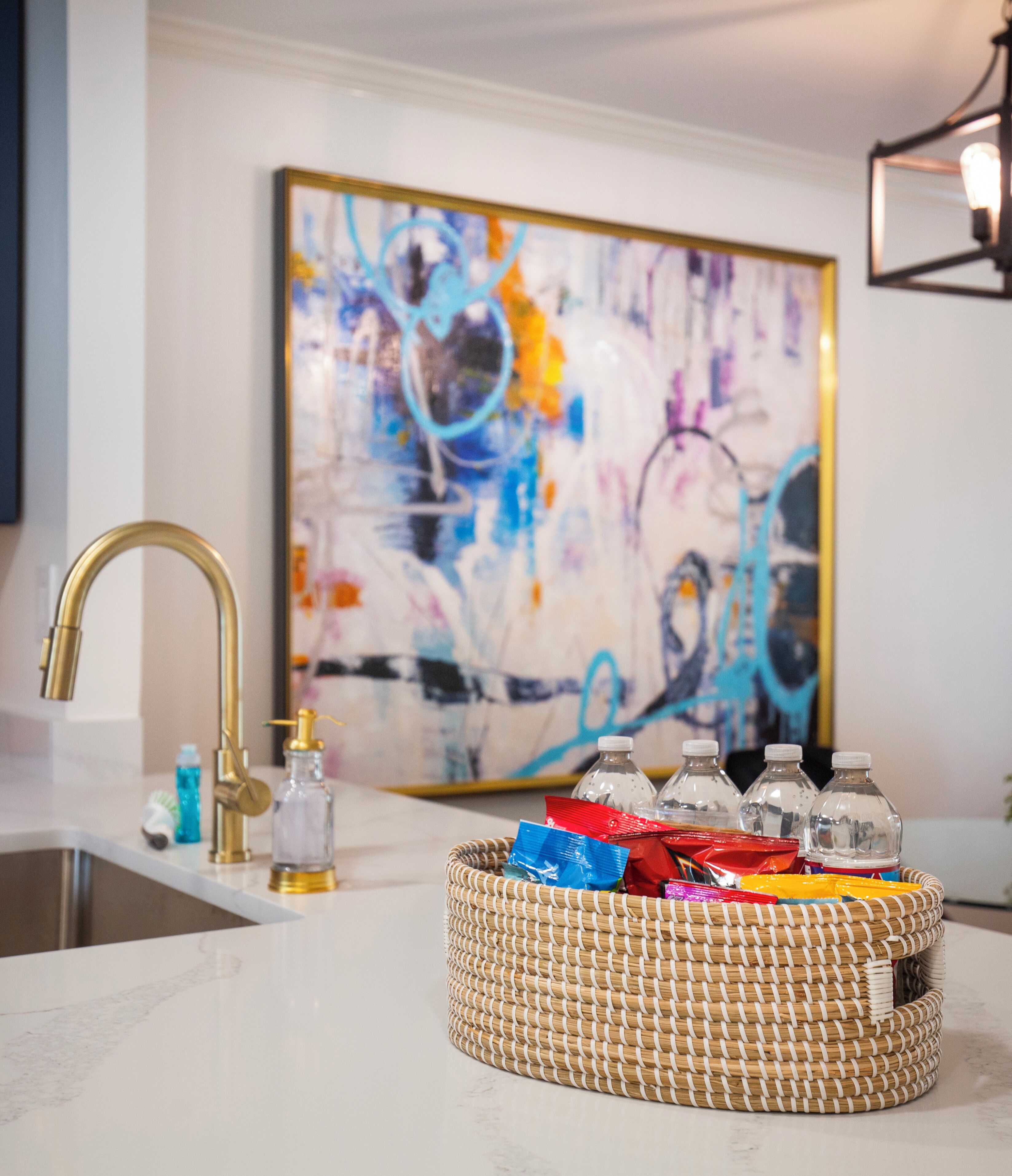 A kitchen with a modern faucet, a woven basket with snacks, and a colorful abstract painting on the wall.