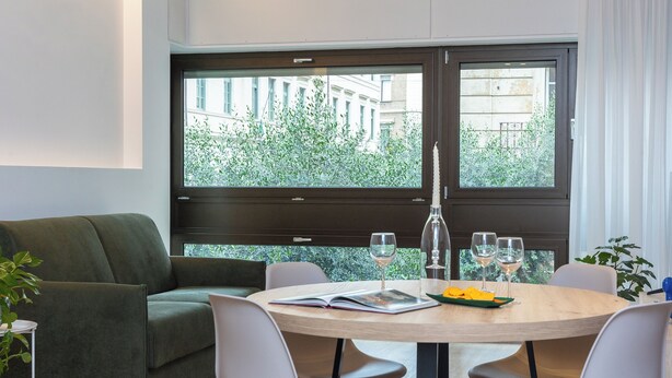 A modern living room with a green sofa, a round wooden dining table with four chairs, and large windows offering a view of greenery and buildings.