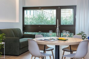 Un salon moderne avec un canapé vert, une table de salle à manger ronde en bois et quatre chaises, ainsi que de grandes fenêtres offrant une vue sur la verdure et les bâtiments.