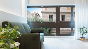A modern living room with a green sofa, a potted plant, and a view through a large sliding glass door to an outdoor balcony.