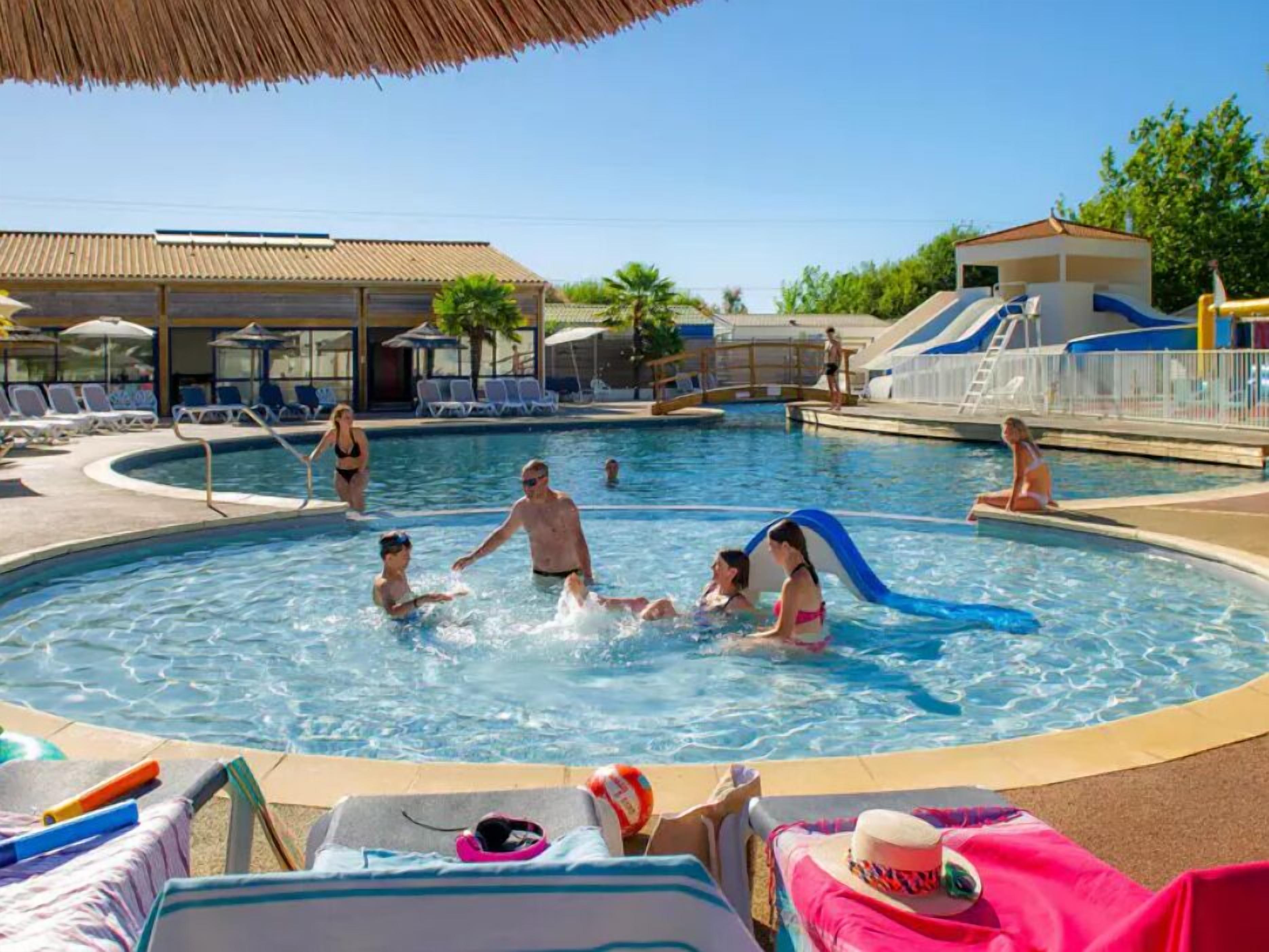 Una piscina con sillas de descanso y sombrillas, un tobogán y un edificio con tejado de tejas.