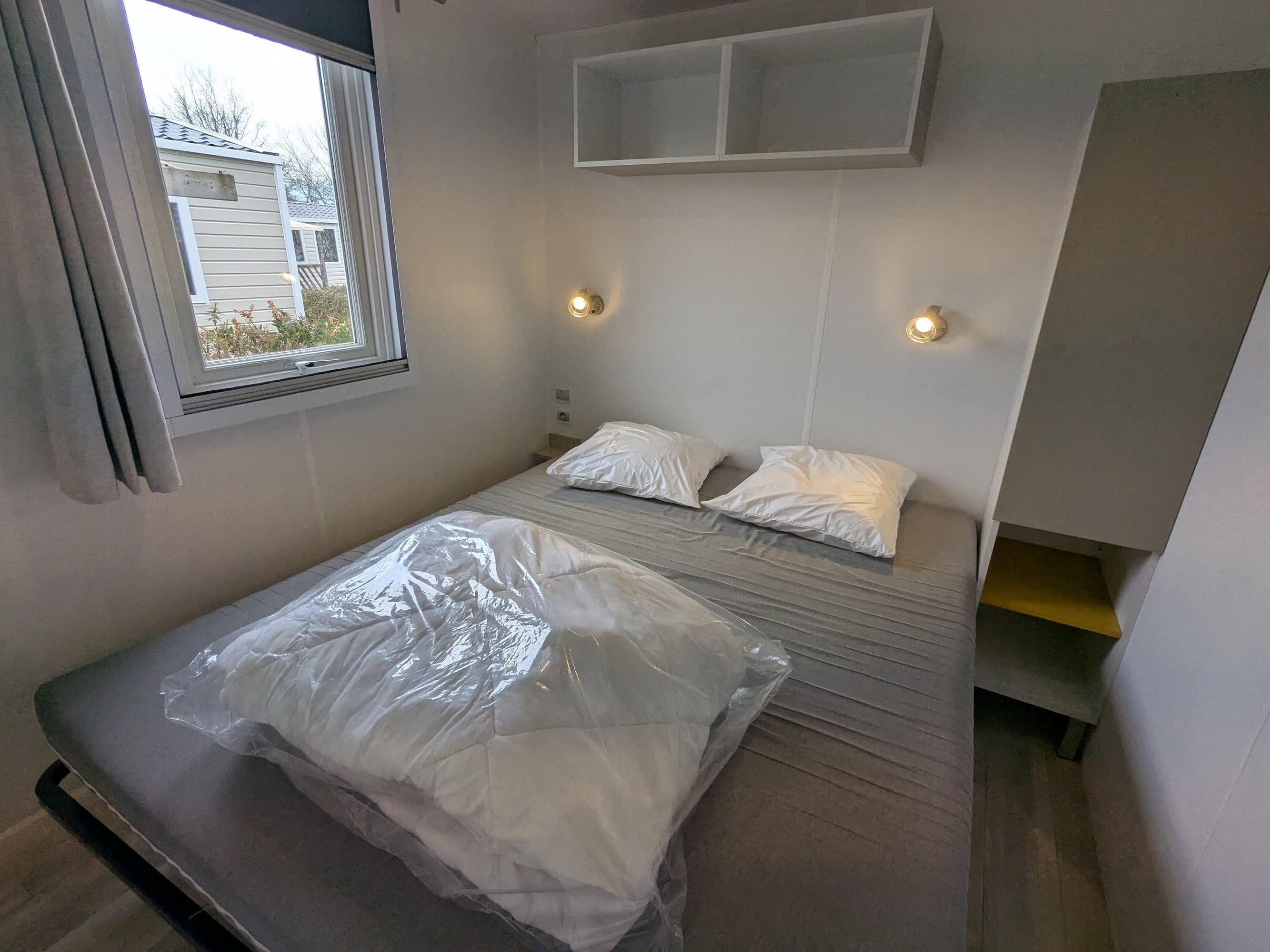 Una habitación pequeña con cama individual, ventana, dos luces fijadas a la pared y un estante empotrado sobre la cama.