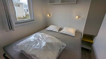 Una habitación pequeña con cama individual, ventana, dos luces fijadas a la pared y un estante empotrado sobre la cama.