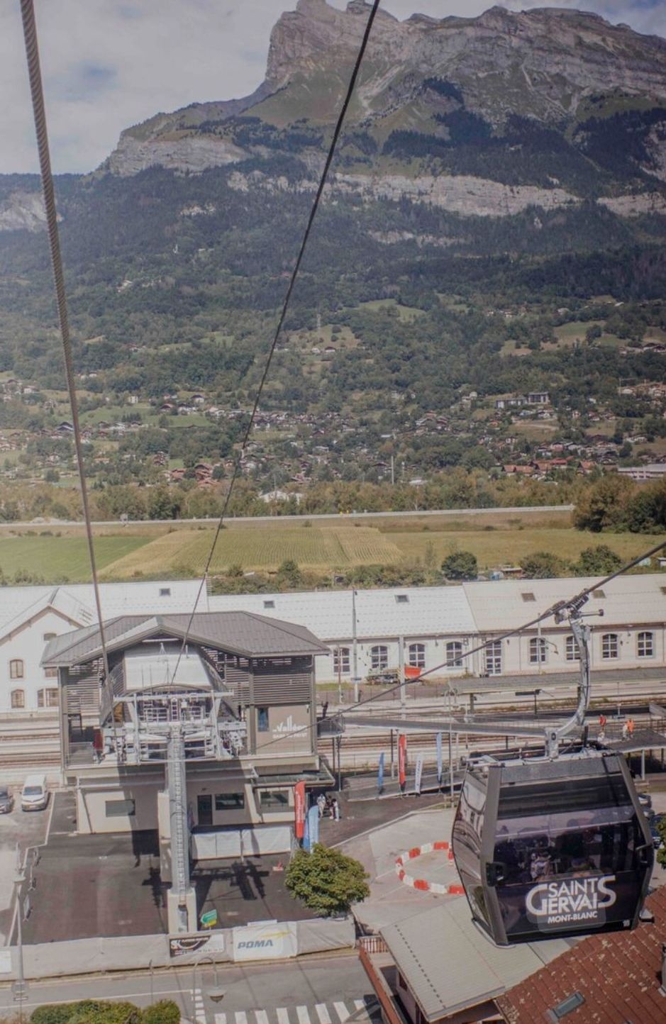 Een kabelbaanstation met een berg op de achtergrond, een gebouw met een bord waarop 'SAINTS GERVAIS MONT BLANC' staat, en een kabelbaan met de tekst 'SAINTS GERVAIS MONT BLANC'.
