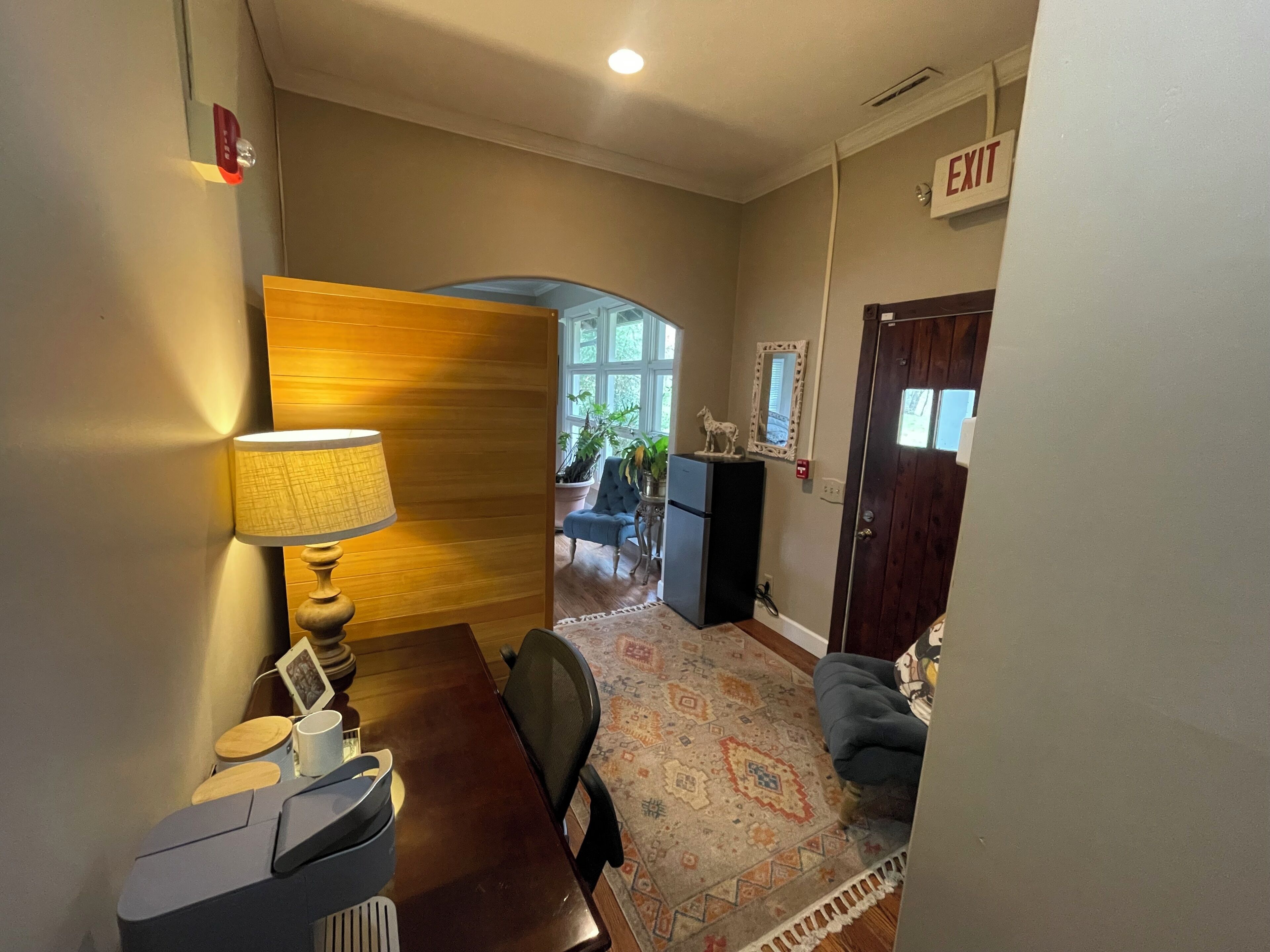 A hallway with a wooden wall, a lamp, a desk, a chair, a rug, and an exit sign.