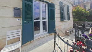 A building with a traditional facade, a set of double doors with green shutters, a white bench, and a small garden area with flowers and a potted plant. - Central Design Flat With Parking  (Bogliasco)
