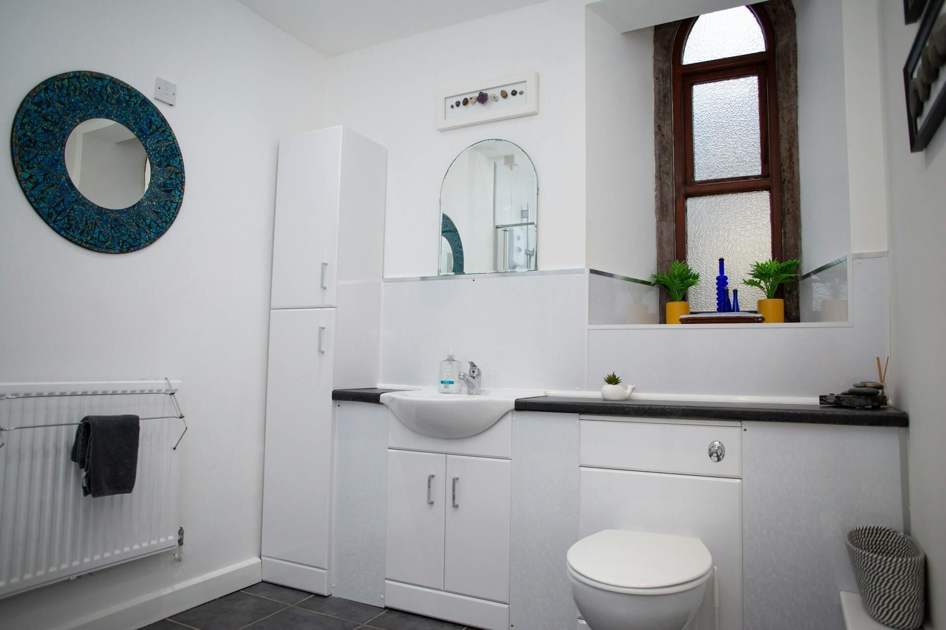 Et moderne badeværelse med en hvid vask, toilet og spejl. Der er et vindue med planter i vindueskarmen og en radiator med et håndklæde hængende på.