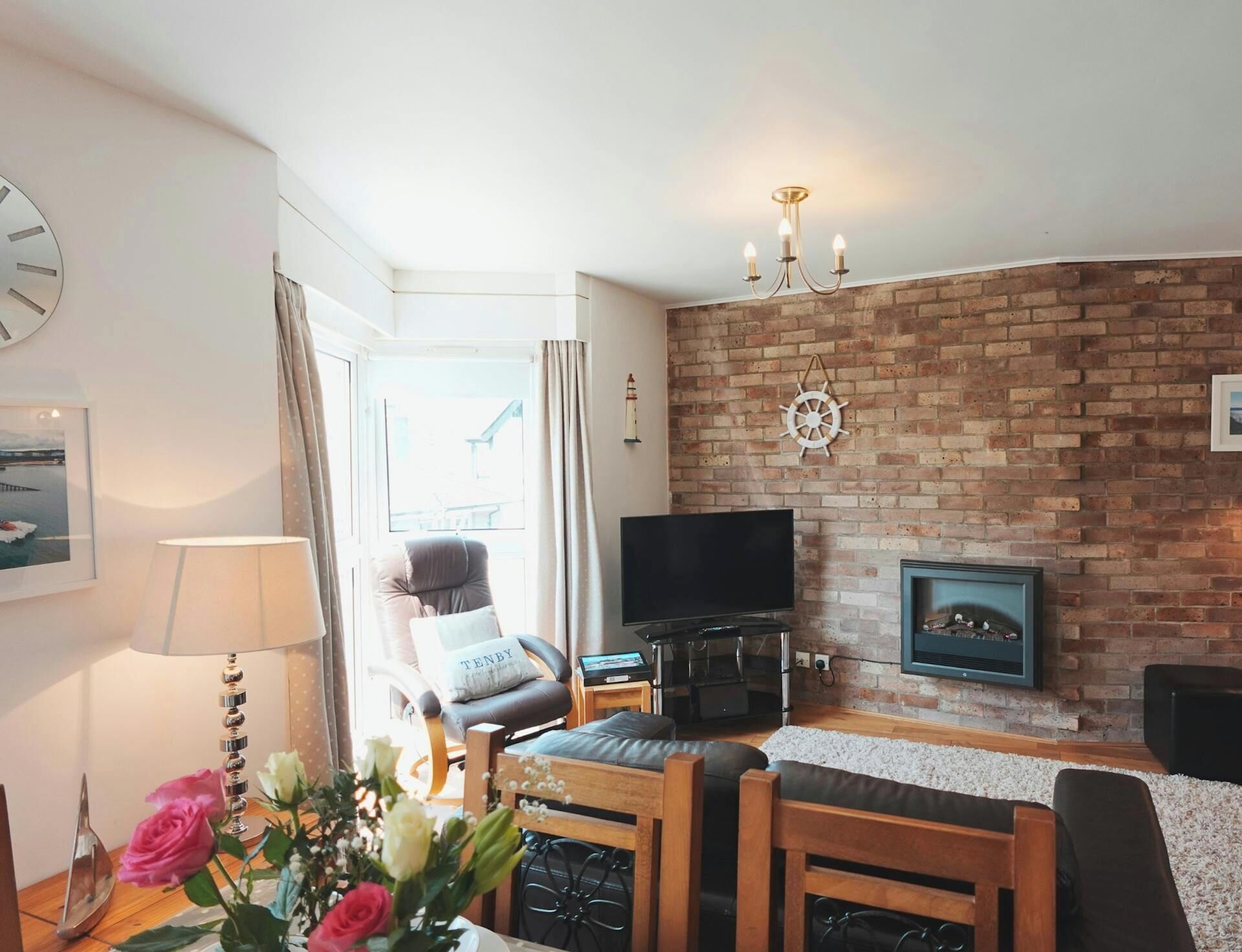 A living room with a brick wall, a flat-screen TV, a fireplace, a wooden coffee table, and a chair with a cushion.