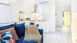 A modern living room with a blue sofa, a beige throw blanket, and a colorful pillow. A kitchen with white cabinets and a microwave is visible in the background.