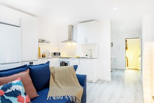 Una sala moderna con un sofá azul, una manta beige y un cojín colorido. Se ve una cocina con gabinetes blancos y un microondas al fondo.