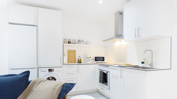 A modern kitchen with white cabinets, a microwave, a stove, and a sink. There is a round white table and a blue sofa with a beige throw blanket.