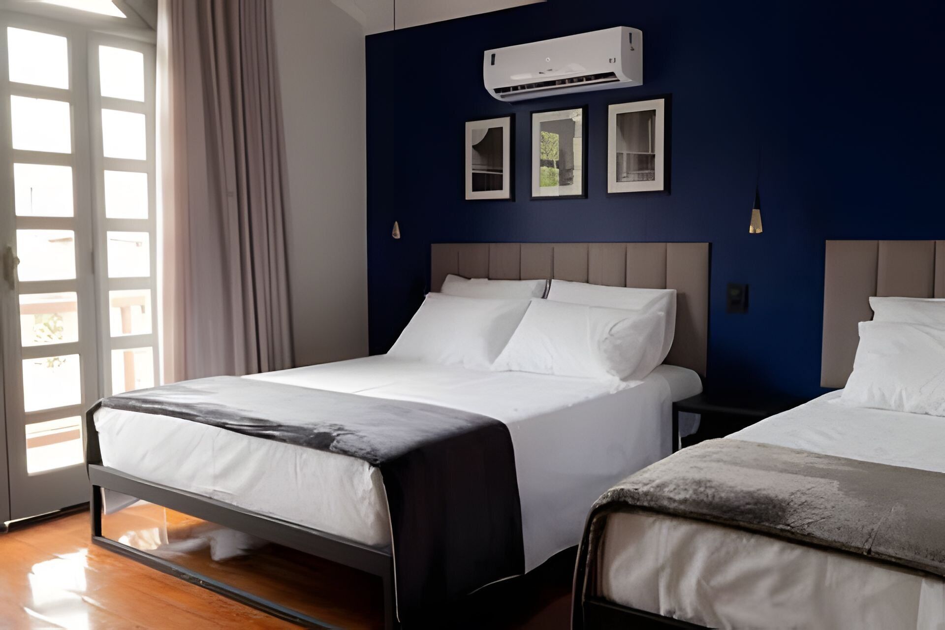 Une chambre d’hôtel avec deux lits, une fenêtre avec des rideaux, un appareil de climatisation et des tableaux encadrés au mur.