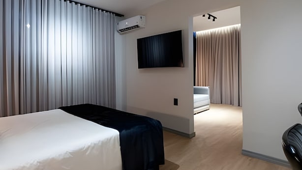 Habitación de hotel moderna con cama, televisor de pantalla plana y zona de estar con sofá y cortinas.