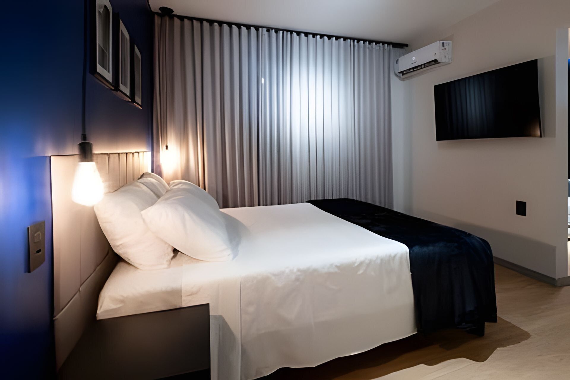 Quarto de hotel moderno com uma cama bem arrumada, uma televisão montada na parede e um aparelho de ar condicionado.