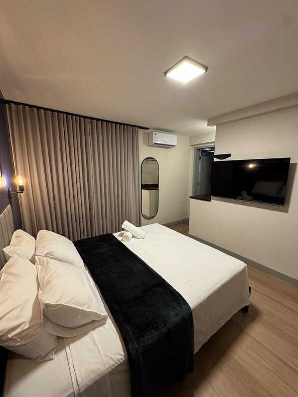 A hotel room with a bed, a flat-screen TV mounted on the wall, an air conditioning unit, and a mirror. - Conde de Linhares Flats (Belo Horizonte)