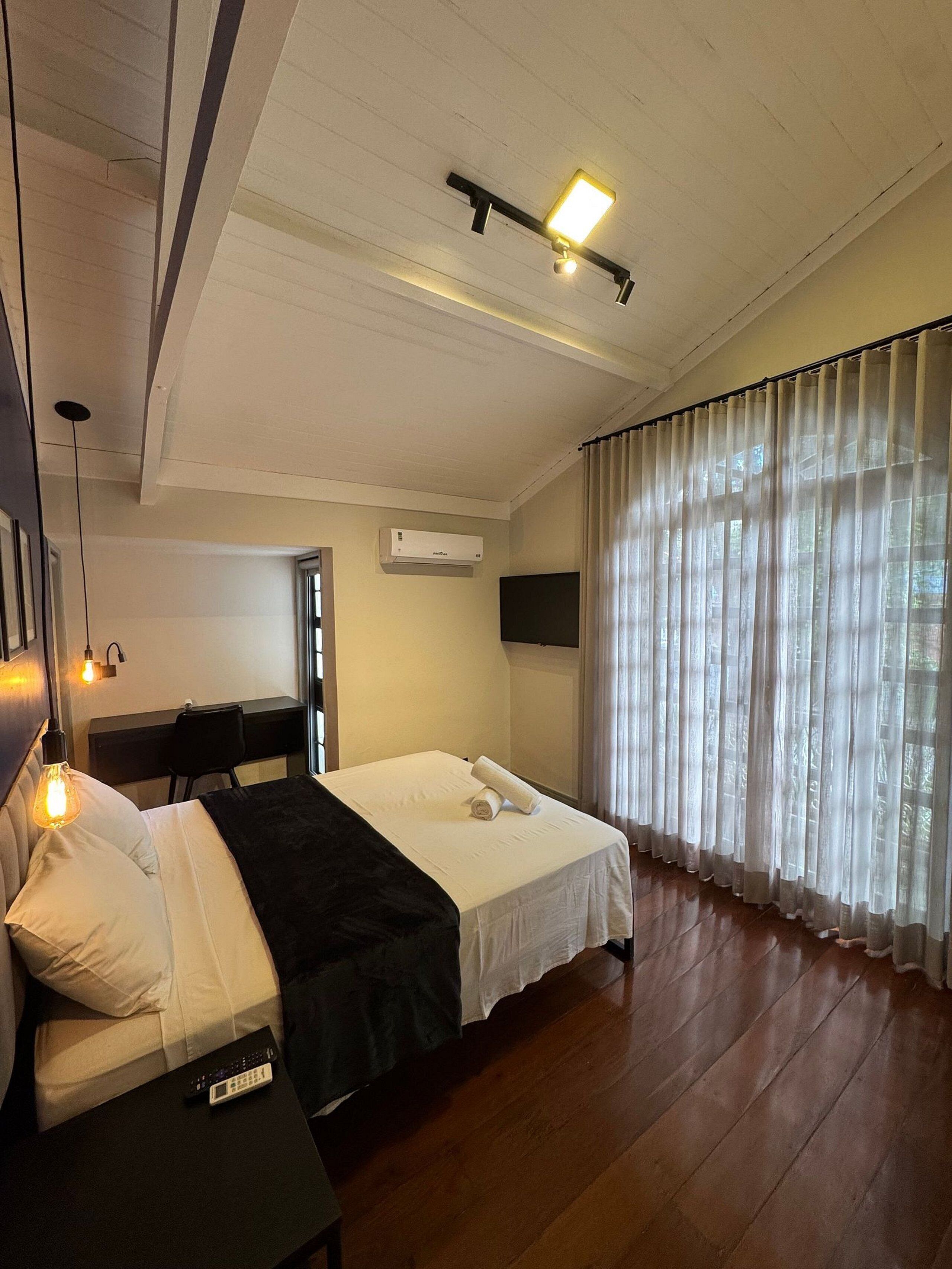 A hotel room with a bed, a desk, an air conditioner, and a window with sheer curtains.