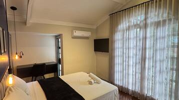 A hotel room with a bed, a desk, an air conditioner, and a window with sheer curtains.