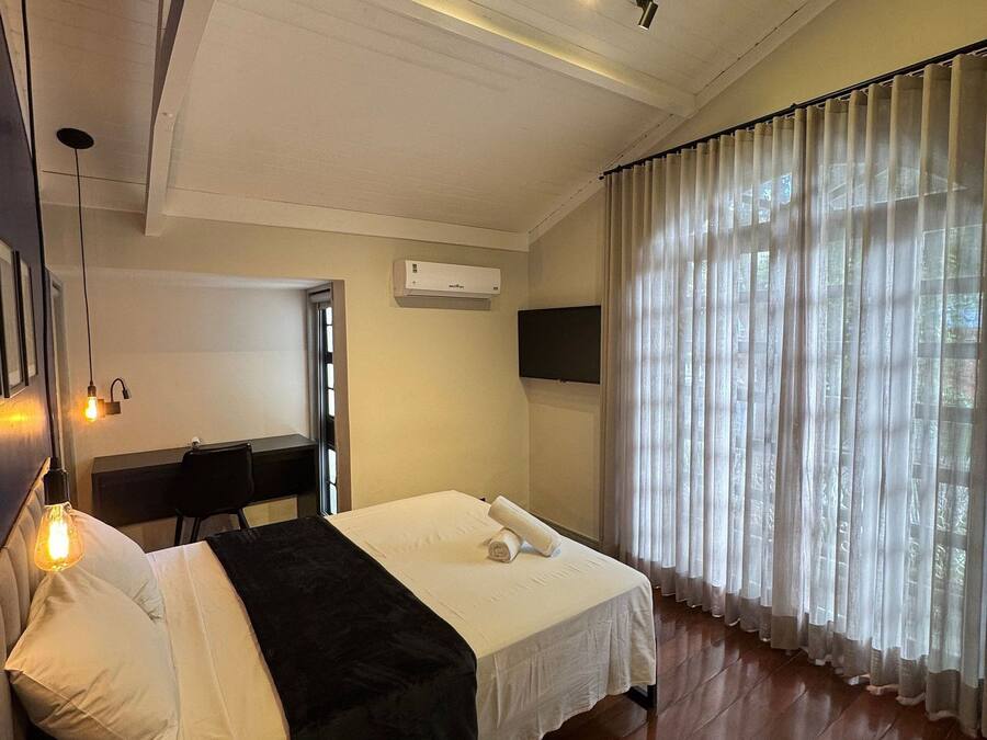 A hotel room with a bed, a desk, an air conditioner, and a window with sheer curtains.