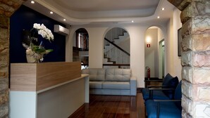 Zona de recepción con un mostrador de madera, un sofá blanco y un sillón azul. Se observa una escalera en el fondo que conduce a un piso superior.