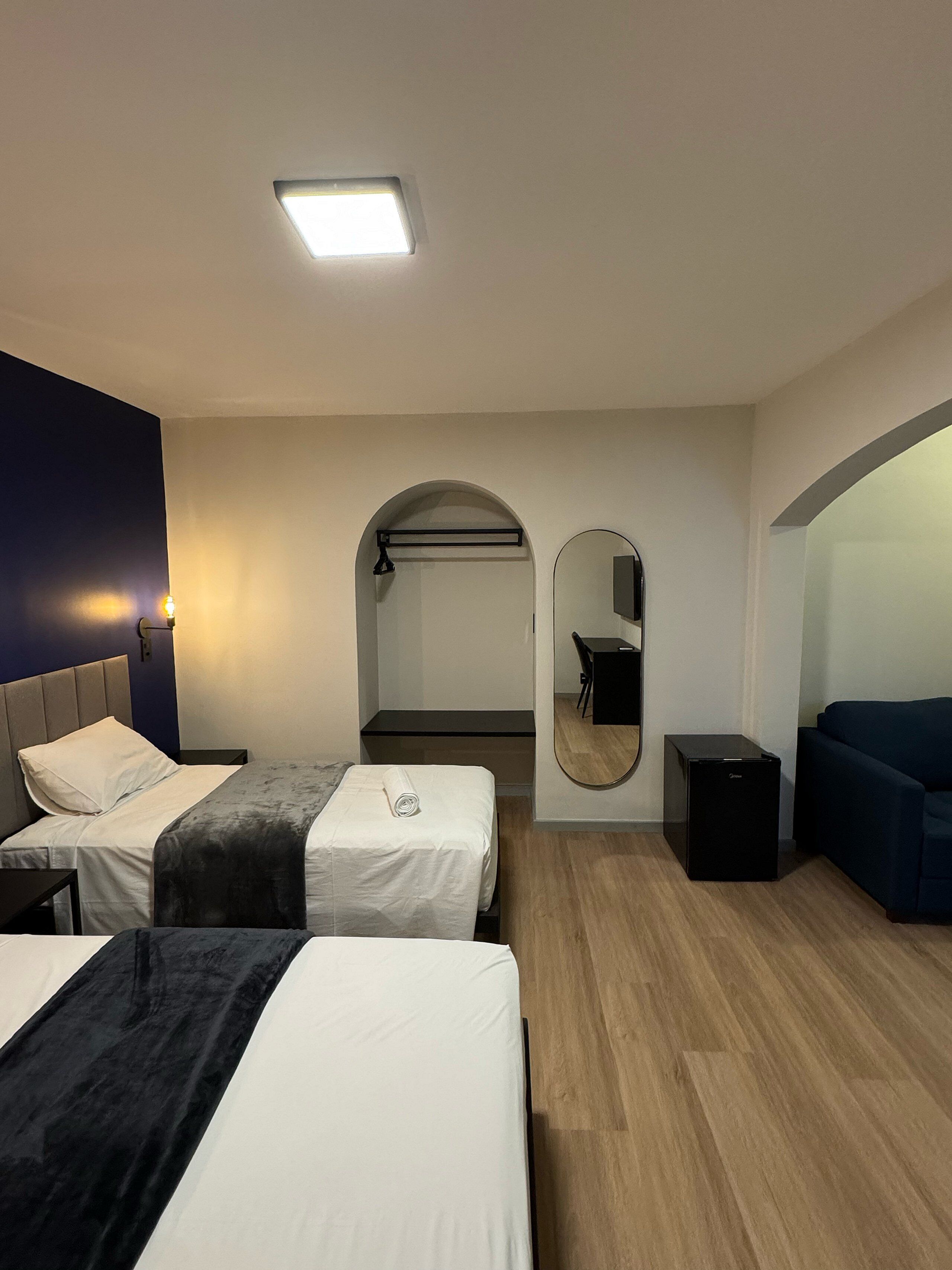 Una habitación de hotel moderna con dos camas, un escritorio pequeño, un espejo y una kitchenette compacta.
