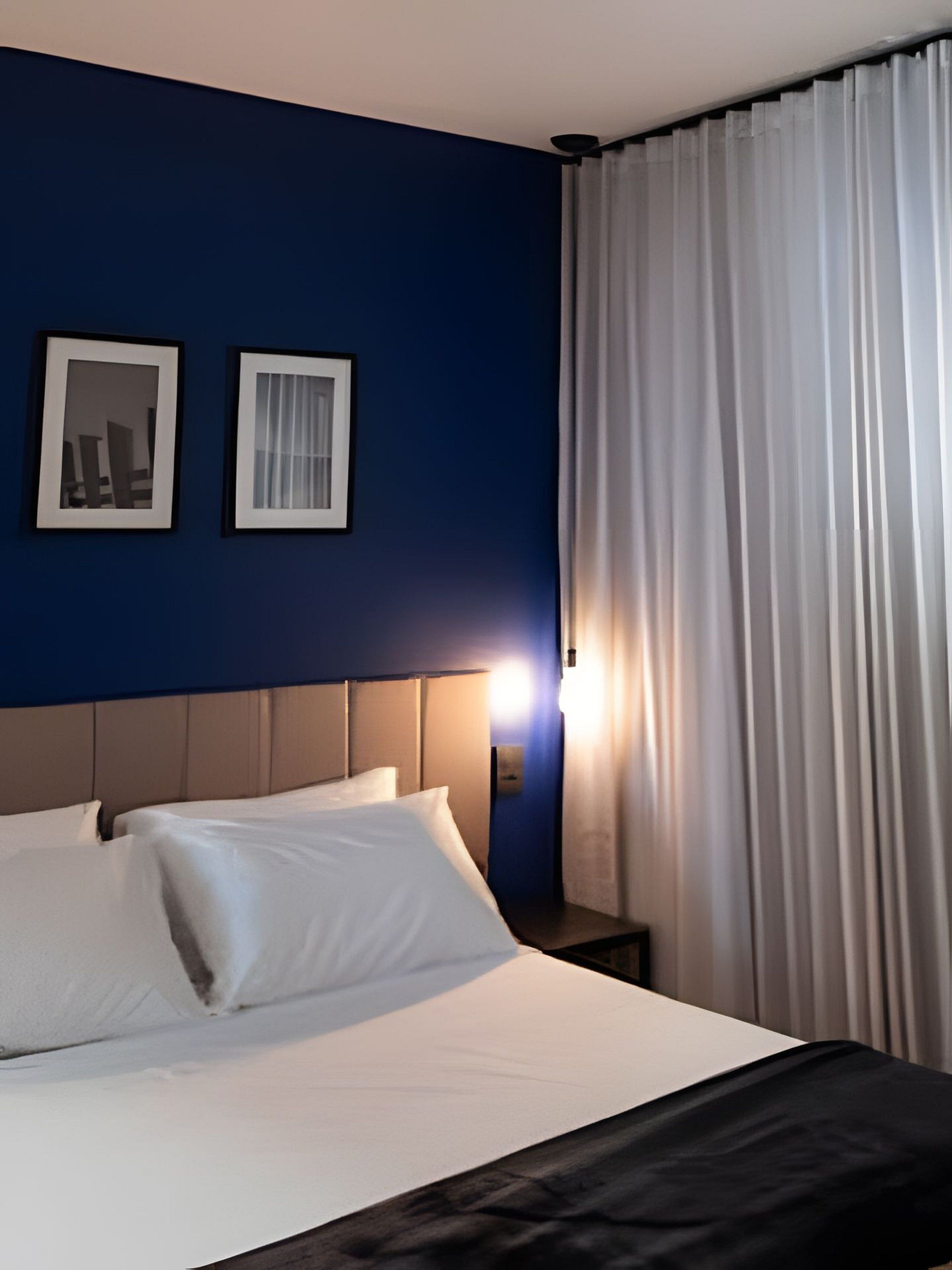 A hotel room with a bed, two framed pictures on the wall, and a window with curtains.
