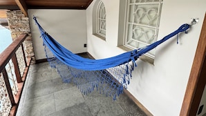 Una hamaca en un balcón con baldosas de piedra, barandilla de madera y ventanas arqueadas.
