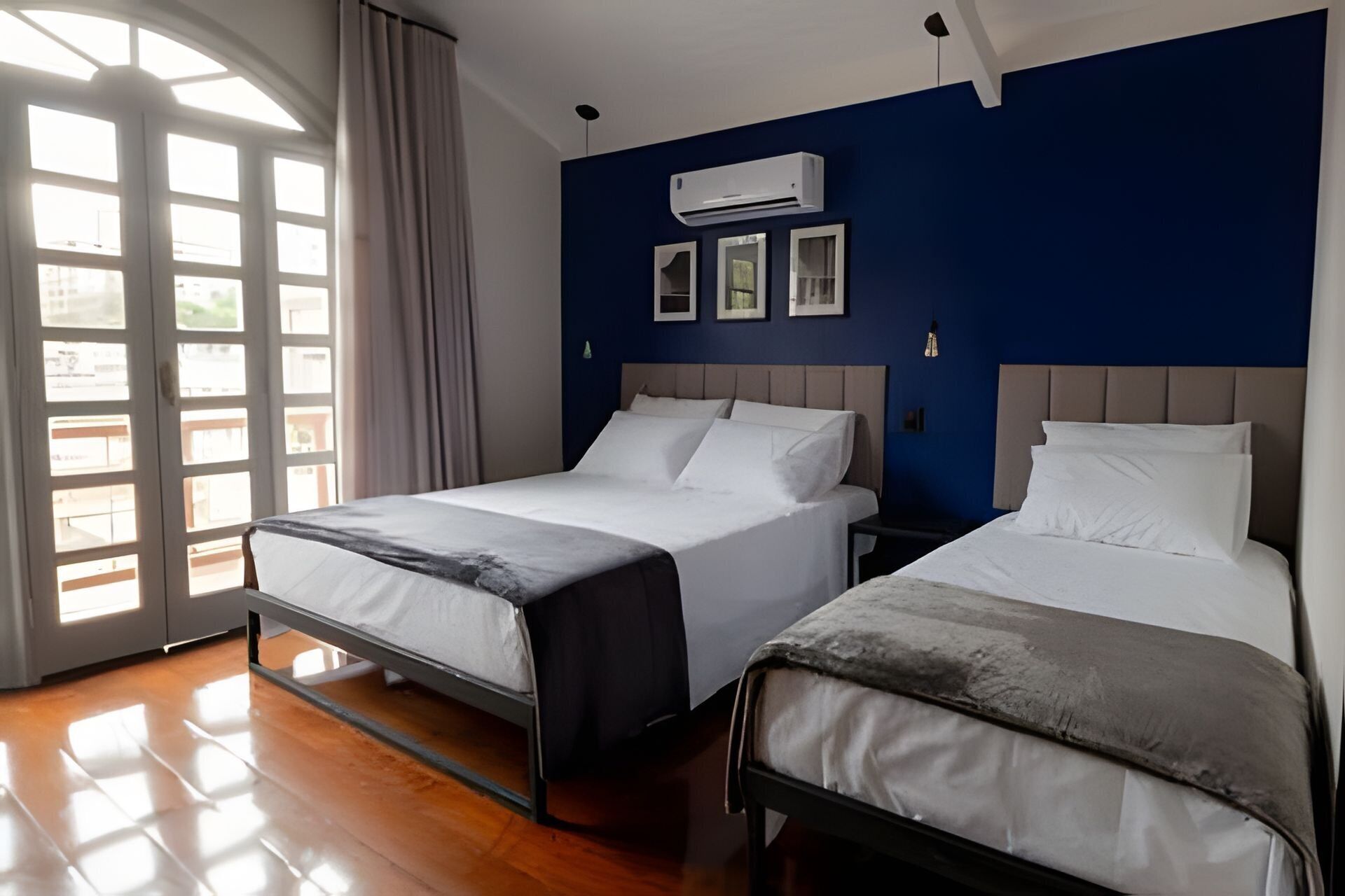 Habitación de hotel con dos camas, un ventanal con cortinas, un equipo de aire acondicionado y cuadros en la pared.