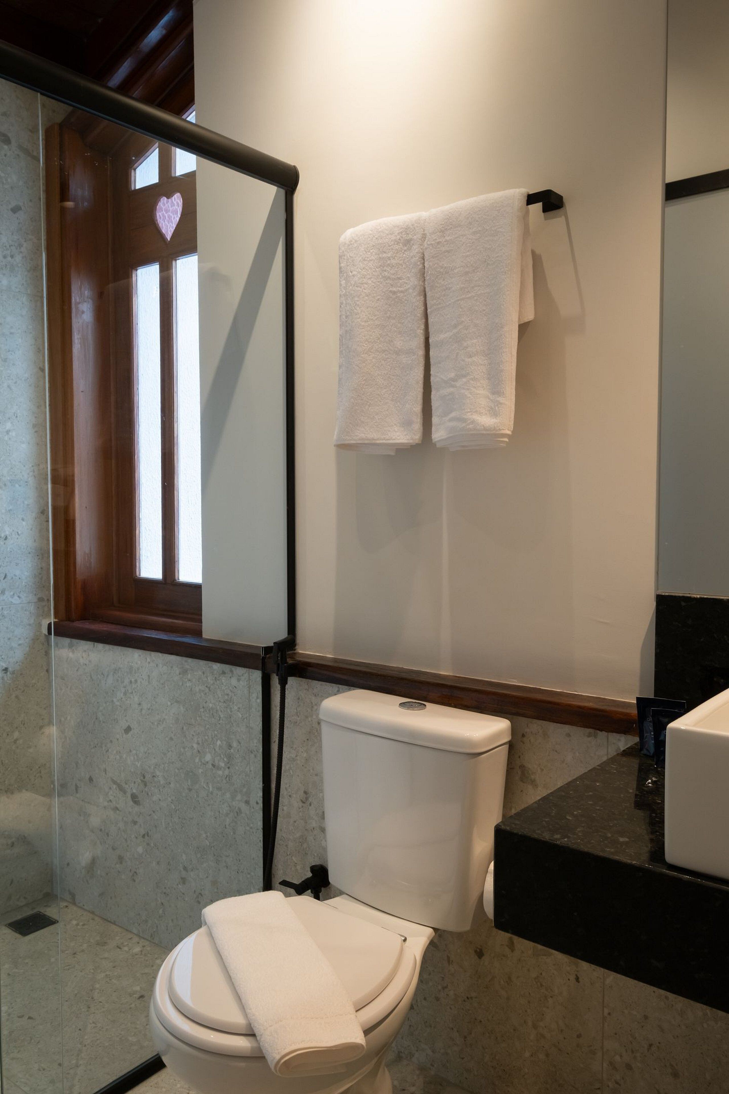 Un baño con inodoro, un perchero con dos toallas, una ventana decorada con un corazón y un espejo.