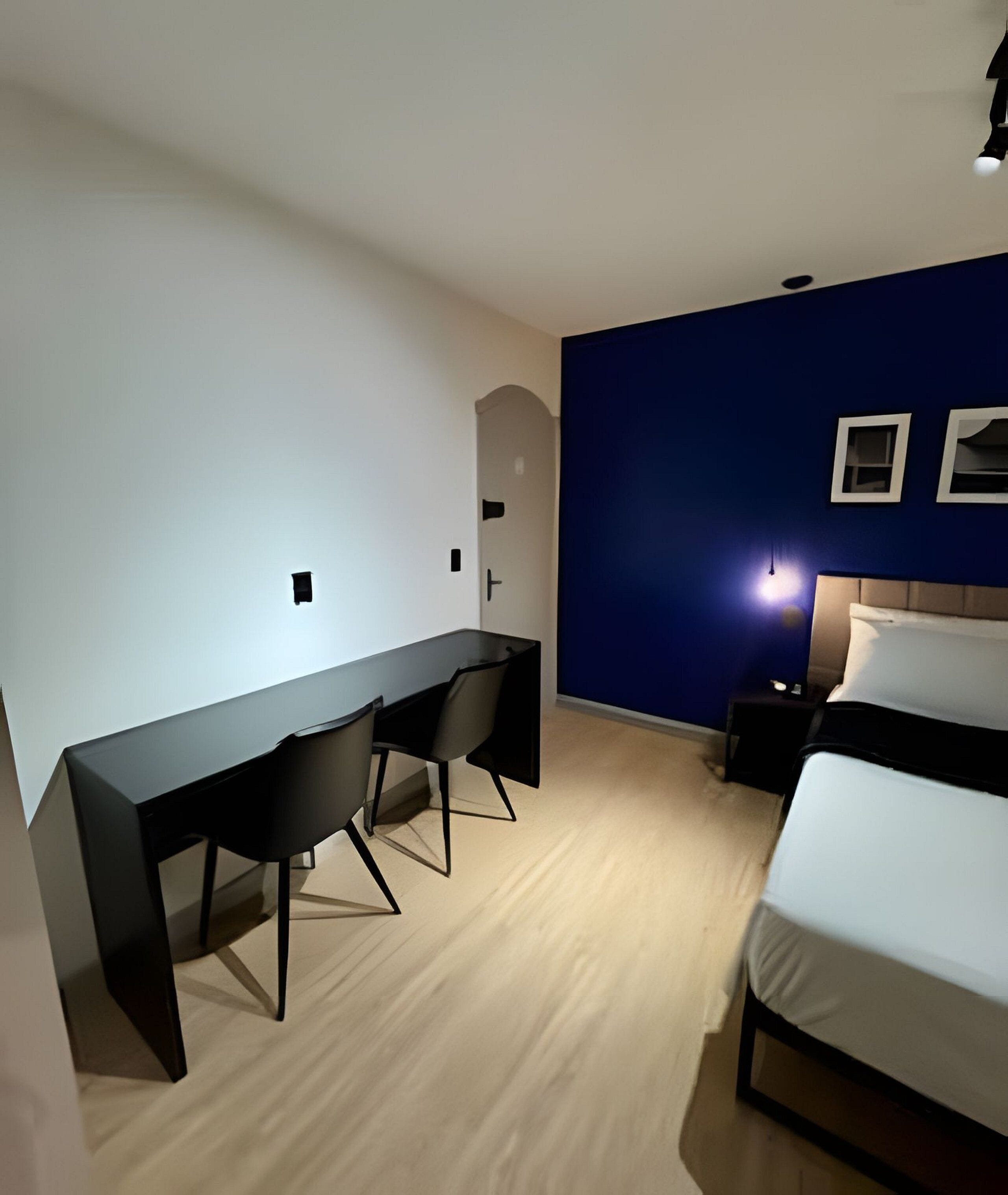 Un dormitorio con una cama, un escritorio con dos sillas, una mesita de noche y una lámpara de pared.