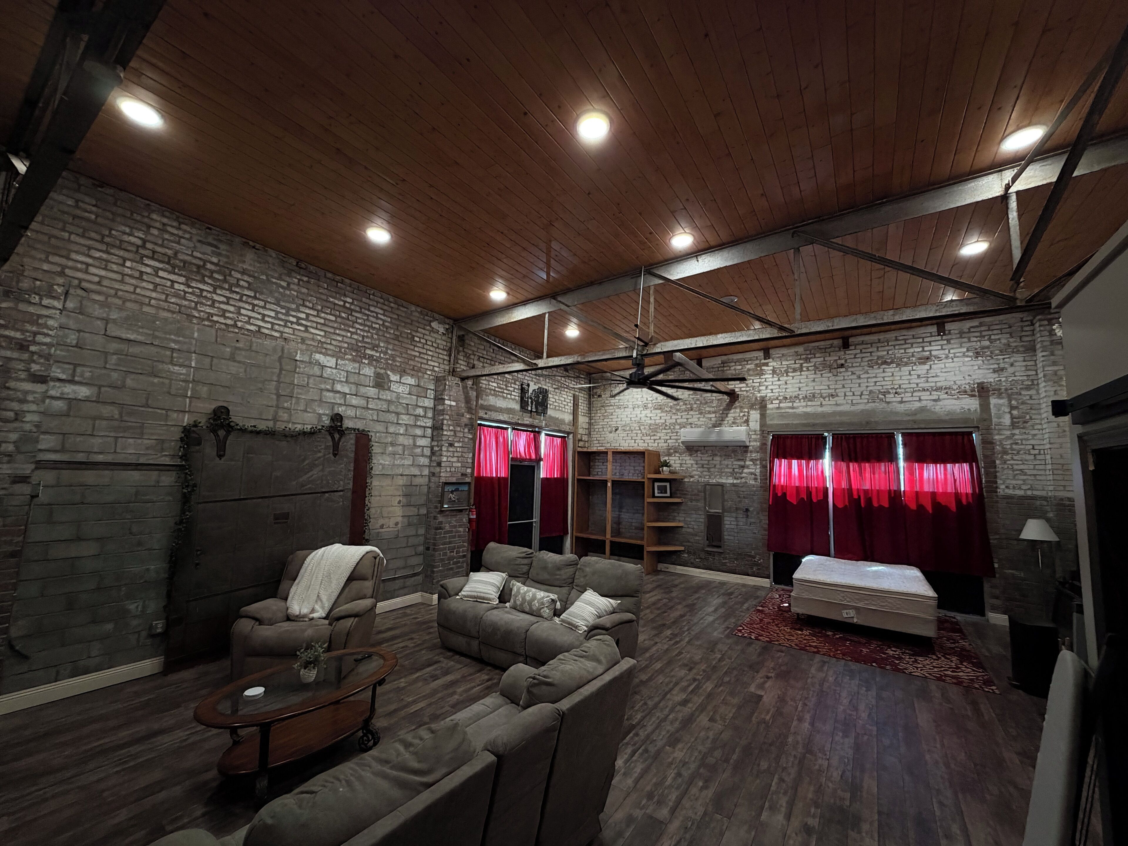 A spacious room with a brick wall, wooden ceiling, and a ceiling fan. There is a sofa, a small table, and a bed with red curtains.