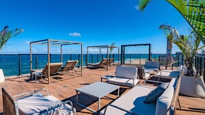 A rooftop terrace with lounge chairs, tables, and a glass balustrade overlooking the ocean.